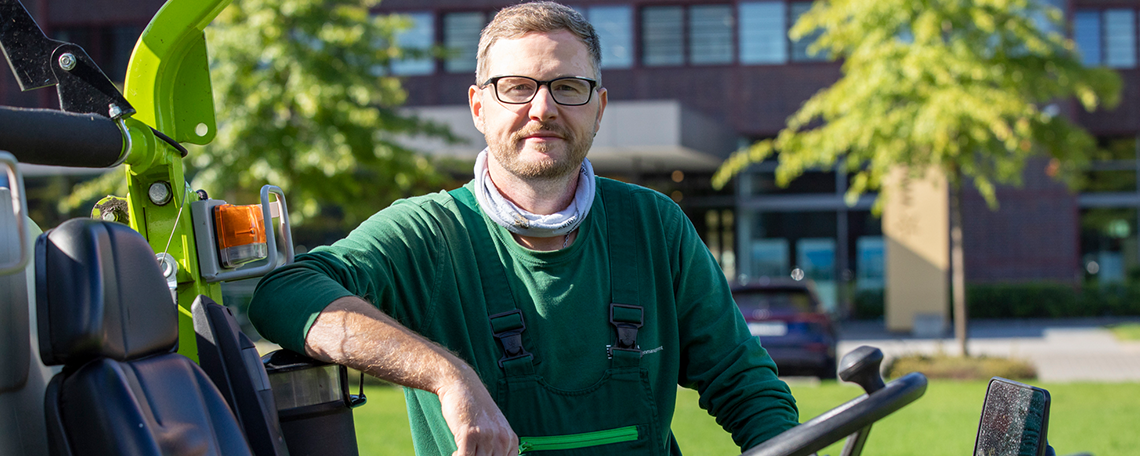 Ein Mann mit kurzen braunen Haaren, Bart und Brille steht neben einem Aufsitzmäher im Freien. Er trägt grüne Arbeitskleidung mit Latzhose und ein helles Halstuch. Im Hintergrund sind Bäume, eine Wiese und ein modernes Gebäude mit großen Fenstern zu sehen. 